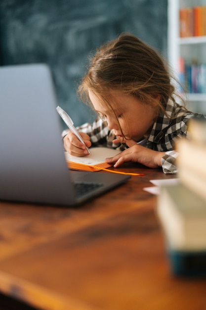 Plan vertical d'une écolière du primaire focalisée faisant ses devoirs en écrivant des notes dans un cahier en papier assis à table avec un ordinateur portable. Adorable élève sérieuse écolière e-learning en ligne à l'aide d'une application informatique.