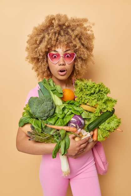 Photo plan vertical d'une femme bouclée étonnée porte des légumes frais préfère des aliments sains porte des lunettes de soleil coeur rose surpris d'avoir une si grande récolte isolée sur fond marron épicerie verte