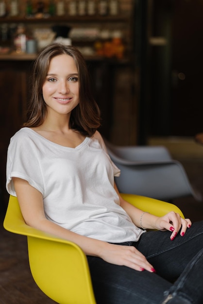 Plan vertical d'une femme souriante a des cheveux noirs sourire à pleines dents vêtu d'un t-shirt blanc est assis dans un fauteuil jaune a une manucure rouge sent des poses de relaxation dans la chambre seule Beauté des gens et temps libre