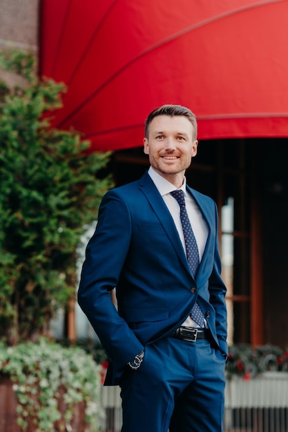 Plan vertical d'un jeune homme séduisant, propriétaire d'une entreprise, célèbre que quelque chose se tient à l'extérieur, garde les mains dans la poche, vêtu d'un costume noir formel, heureux de parler avec quelqu'un pour discuter du travail ensemble