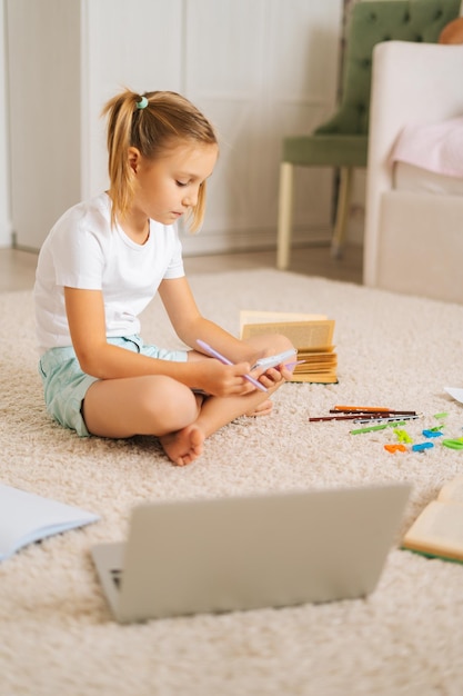 Plan vertical d'une jolie fille primaire concentrée écrivant ses devoirs dans un cahier assis sur le sol à la maison