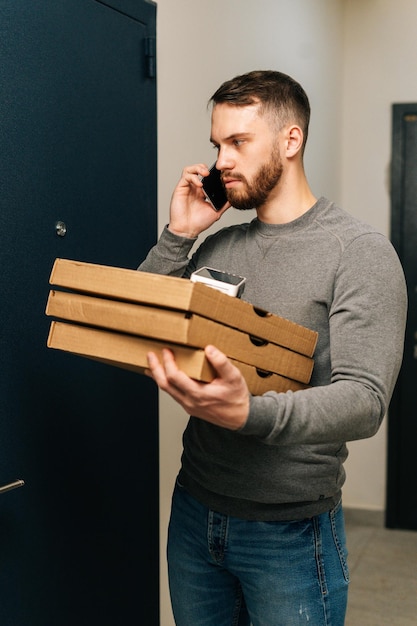 Plan vertical d'un livreur debout près de la porte de l'appartement du client avec des boîtes de pizza chaude et appelant le client au téléphone