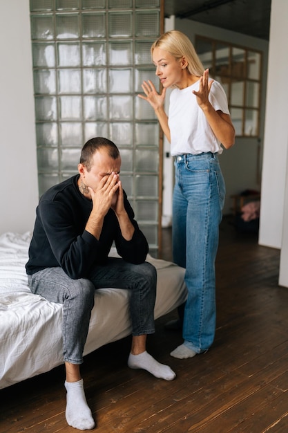 Plan vertical d'un mari caucasien frustré assis sur son lit, ignorant une femme blonde debout en colère, arguant de blâmer un homme bouleversé de problèmes à la maison