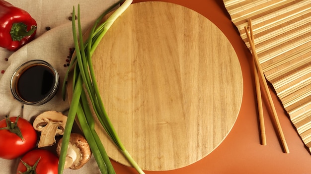 Planche de bois ronde, sauce et divers légumes sur la table, bâton en bois sur fond marron. Mise à plat. Copier l'espace