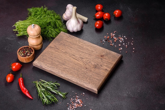 Une planche à découper en bois avec des épices, des herbes, des tomates cerises et du sel sur un fond de béton noir
