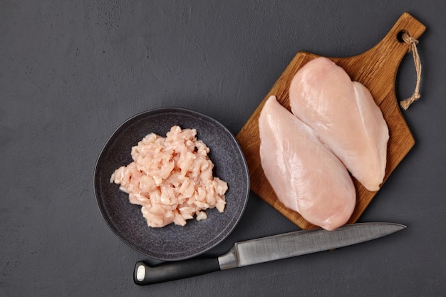 Planche à découper en bois marron avec des morceaux de filet de poitrine de poulet et un bol de viande de volaille hachée sur une table en béton foncé Vue de dessus avec copie espace à plat