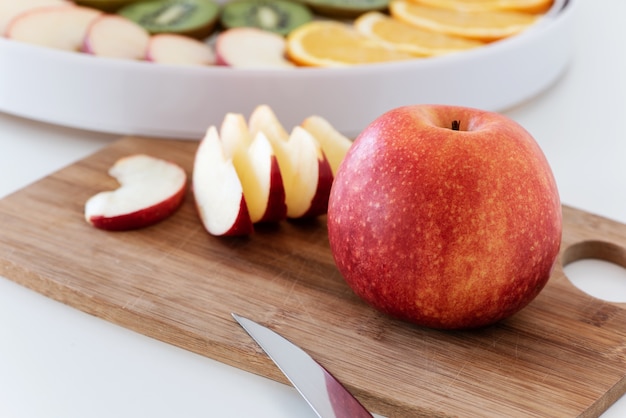 Planche à découper avec un couteau, pomme rouge et tranches de pomme. Derrière se trouve un déshydrateur avec des tranches d'orange, de kiwi et de pommes.