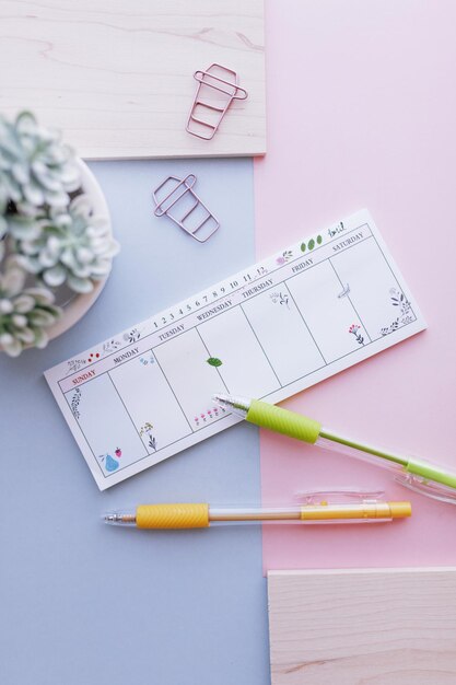 Photo planificateur de sept jours sur fond rose et bleu avec des stylos et des pinces à papier