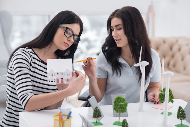 Planification innovante. Pensive femme handicapée et collègue posant à table et travaillant avec des modèles d'énergie alternative