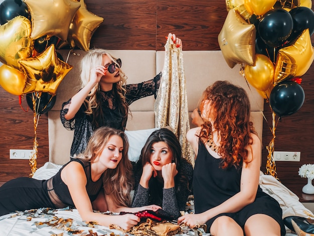 Plans de soirée entre filles. Temps libre pour faire la fête. Les femmes allongées sur le lit en choisissant une tenue de paillettes. Confettis scintillants et ballons autour.