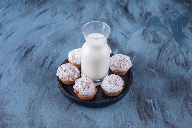 Plaque noire avec cupcakes crémeux sucrés et verre de lait sur une surface en marbre.