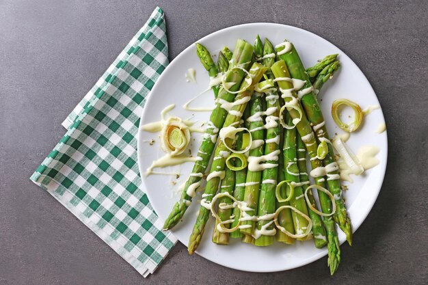 Plat d'asperges fraîches avec des tranches d'oignon sur une assiette blanche avec une serviette en coton à carreaux sur une table grise