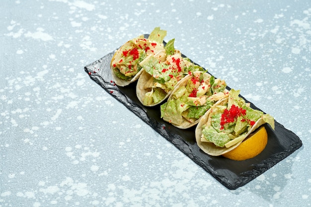 Plat de cuisine mexicaine - tacos au saumon, laitue, sauce blanche et caviar tobiko sur une plaque noire sur fond bleu