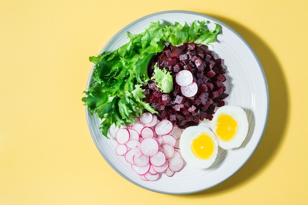 Un plat diététique à base de légumes. Tartare de betterave, radis, salade de frise et œuf à la coque sur une assiette sur une table jaune. Vue de dessus. Espace de copie