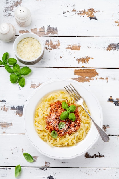 Plat italien traditionnel de pâtes fettuccine avec sauce bolognaise, basilic et parmesan dans une assiette blanche sur fond de bois clair. Vue de dessus.