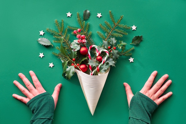 Plat de Noël créatif posé sur du papier vert. Mains et décorations de Noël en cône de placage - brindilles de sapin et de houx, boules, cannes de bonbon, étoiles en bois et fruits rouges.