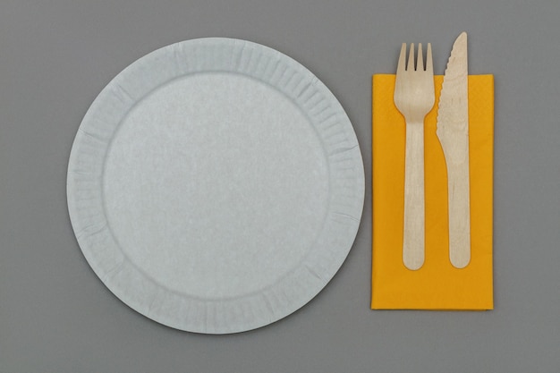 Plat en papier blanc, fourchette en bois et couteau sur serviette orange sur surface grise, vue du dessus.