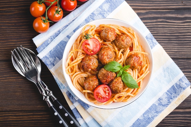 Plat traditionnel italo-américain spaghetti aux boulettes de viande, sauce tomate et basilic dans un bol, avec couverts et serviette, fond en bois rustique