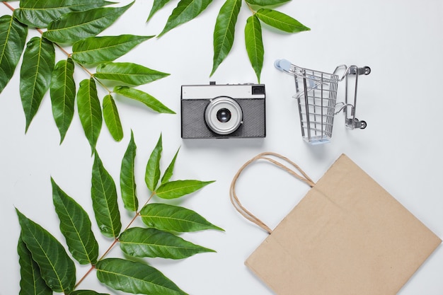 Photo plat la vie toujours accro au shopping. panier, sac en papier écologique, appareil photo rétro sur fond blanc parmi les feuilles vertes.