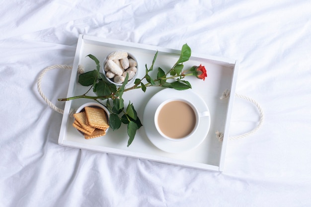 Plateau blanc avec café, biscuits et fleurs sur un lit blanc, vue de dessus.