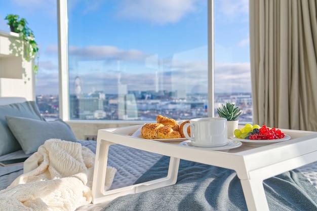 Plateau blanc avec petit-déjeuner savoureux sur le lit dans une chambre lumineuse avec de grandes fenêtres dans un style de vie de gratte-ciel