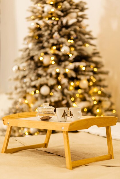 Un plateau en bois se dresse sur le lit sur fond de lumières de Noël Sur le plateau se trouve un pot de biscuits et deux tasses