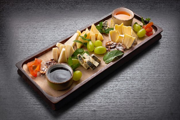 Plateau de fromages sur une planche en bois avec raisins, menthe, miel et confiture, sur une table sombre