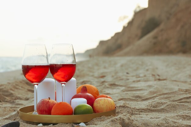 Plateau avec verres de vin, fruits et bougies sur la plage de sable de la mer