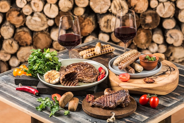 Photo plats de barbecue à base de steaks de bœuf, porc, saucisses de poulet, tomates et piments, vin rouge dans des verres sur fond de barbecue avec du bois de chauffage. vue latérale avec un espace de copie.