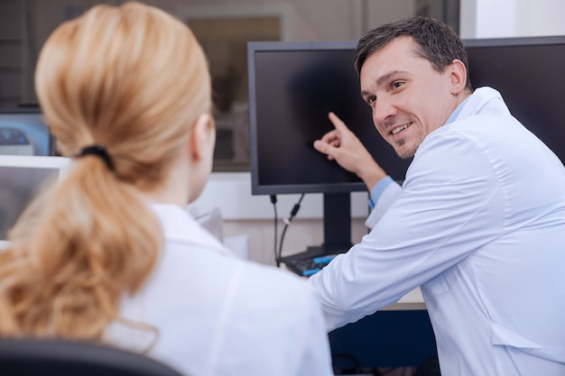 Pleasant happy nice docteur souriant et pointant vers le moniteur tout en travaillant avec son collègue