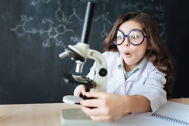 Plein d'émotions positives. Habile fille positive émerveillée assis dans le laboratoire et bénéficiant d'un cours de chimie tout en participant à l'expérience scientifique et à l'aide d'un microscope