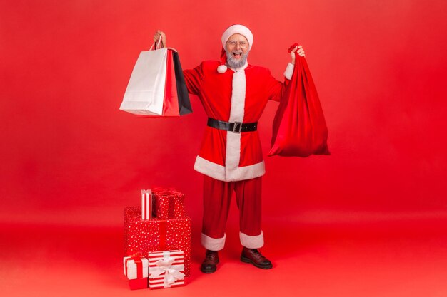 Pleine longueur heureux père noël satisfait tenant et montrant des sacs en papier et un grand sac rouge plein de cadeaux coffrets cadeaux debout sur le sol vacances d'hiver Studio intérieur tourné isolé sur fond rouge