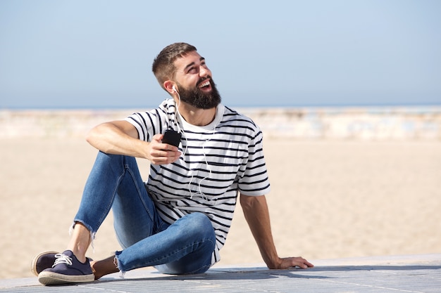 Pleine longueur homme heureux dans les écouteurs assis sur la plage avec un téléphone mobile