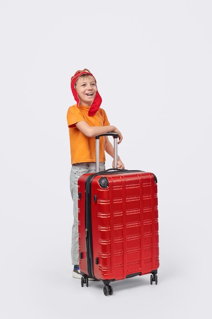 Pleine longueur joyeux petit garçon au chapeau d'aviateur souriant et regardant loin tout en se tenant derrière la valise pendant le voyage sur fond blanc