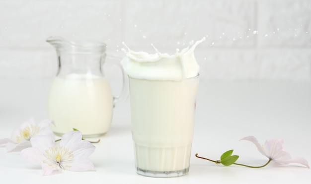 Une pleine tasse en verre de lait frais avec des éclaboussures se dresse sur une table blanche, petit-déjeuner sain