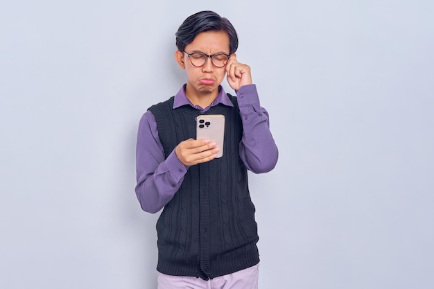 Pleurer jeune homme asiatique en chemise décontractée et gilet à l'aide d'un téléphone portable isolé sur fond blanc Concept de style de vie des gens