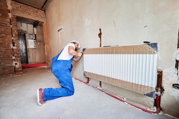 Plombier en combinaison de travail utilisant une clé anglaise lors de l'installation d'un radiateur de chauffage dans une pièce Homme en casque installant un système de chauffages dans un appartement Concept d'installation de radiateurs travaux de plomberie rénovation de la maison