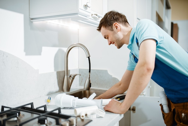 Plombier masculin en problème de fixation uniforme avec robinet dans la cuisine. Homme à tout faire avec évier de réparation de sac à outils, service d'équipement sanitaire à domicile