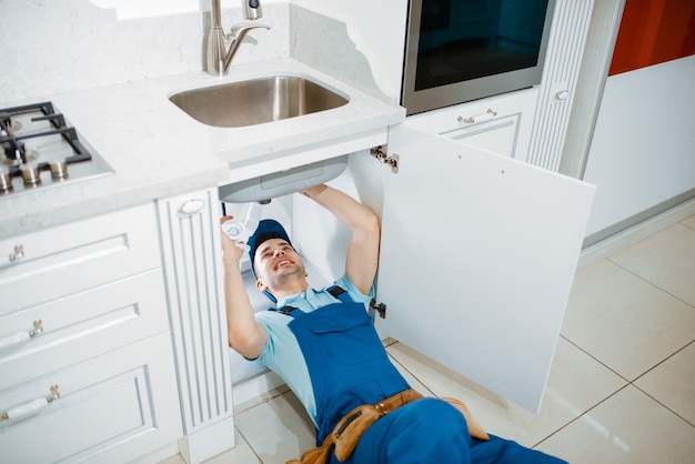 Plombier masculin en uniforme installant un tuyau d'évacuation dans la cuisine. Femme à tout faire avec évier de réparation de sac à outils, service d'équipement sanitaire à domicile