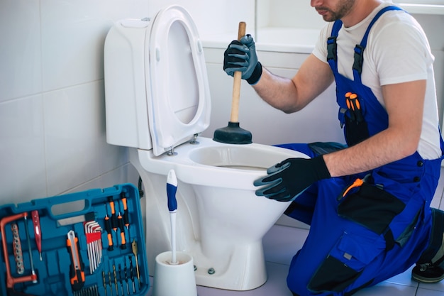 Un plombier professionnel en uniforme répare la cuvette des toilettes dans la salle de bain domestique à l'aide d'outils