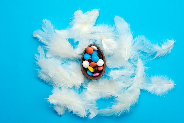 Les plumes blanches reposent sur le concept bleu de Pâques. Bonbons multicolores dans l'oeuf au chocolat