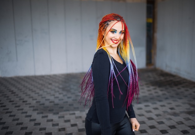 La plus belle fille vêtue d'une robe noire élégante avec un maquillage brillant et des tresses artificielles multicolores. Magnifiquement encerclant et agitant de longues tresses dans le contexte d'un vieux bâtiment.