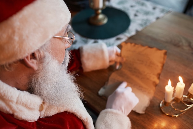 Plus D'épaule Du Père Noël Dans Des Verres Assis à Table Et Lisant Une Lettre Aux Chandelles