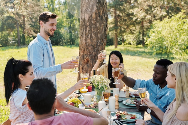 Plusieurs amis internationaux reposants vont tinter avec des verres de vin tout en grillage sur une table de fête servie lors d'un dîner en plein air sous un pin