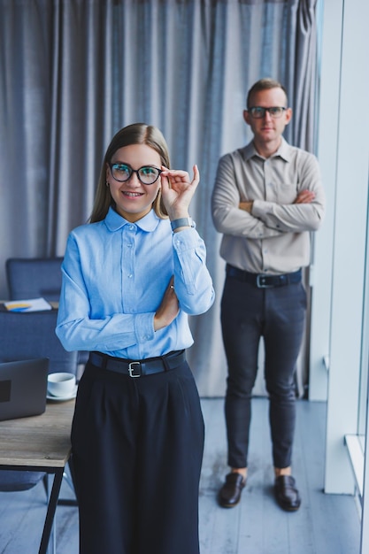 Plusieurs collègues amicaux se tiennent près de la fenêtre du bureau Une femme d'affaires à lunettes se tient sur le fond d'un homme en chemise Mise au point sélective Portrait d'hommes d'affaires