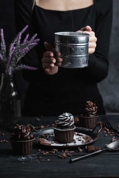 Plusieurs muffins ou cupcakes avec de la crème en forme de chocolat à table noire. La main d'une femme émiette du sucre en poudre sur un gâteau.