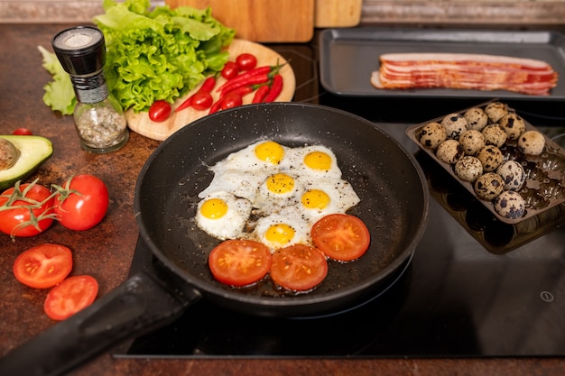 Plusieurs œufs de caille frits et tranches de tomates saupoudrées de poivre noir moulu sur une poêle entourée de légumes frais