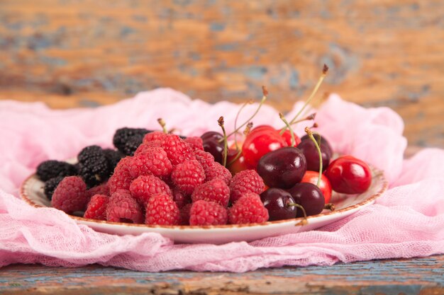 Plusieurs sortes de baies mûres et juteuses sur une assiette. Fond en bois Framboise, cerise, mûre