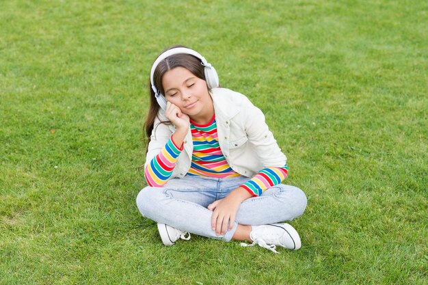 Podcast éducatif Make It Happen Écouter de la musique tout en se relaxant en plein air Enfant fille profiter de la musique prairie d'herbe verte Moment agréable Casque enfant écouter de la musique Casque fille cool écouter de la musique