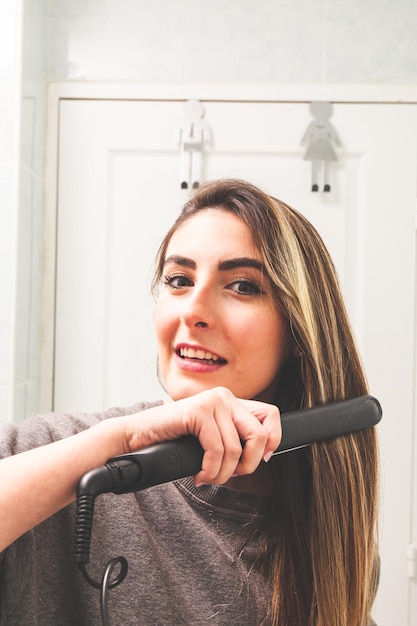 Point de vue miroir d'une jeune femme caucasienne lissant ses cheveux avec un lisseur dans la salle de bain.